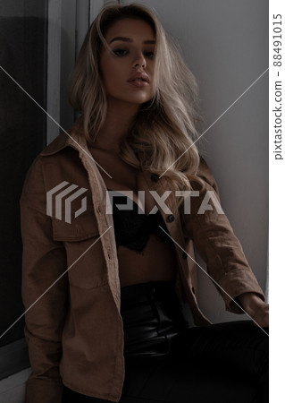 Young gorgeous stylish woman with a beautiful lush breasts in a stylish beautiful lace bra in fashionable leather pants in a beige shirt sitting indoors near the window. Sexy blonde girl. 88491015
