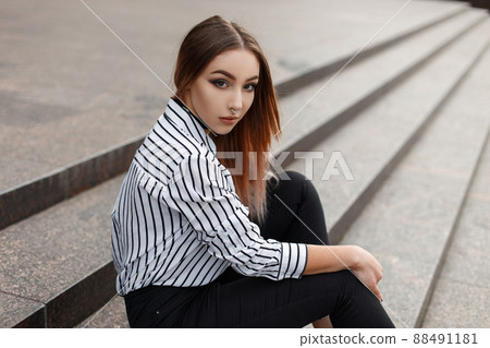 Pretty American young woman in fashionable jeans in a vintage black and white striped blouse with elegant velvet necklaces sits on the vintage steps in the city on a warm spring day. Beautiful girl. 88491181