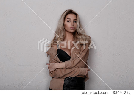 Elegant stylish attractive young woman with beautiful magnificent breasts in a lace bra in vintage beige stylish shirt in leather black pants in a room near a white wall. Sexy blonde girl. 88491232
