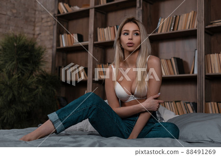 Pretty beautiful young blond woman in a stylish white bra in fashionable blue jeans sitting on the bed near wall with books. Sexy girl model in trendy lingerie resting. Pretty beautiful young blond woman in a stylish white bra in fashionable blue jeans sitting on the bed near wall with books. Sexy girl model in trendy lingerie resting. 88491269
