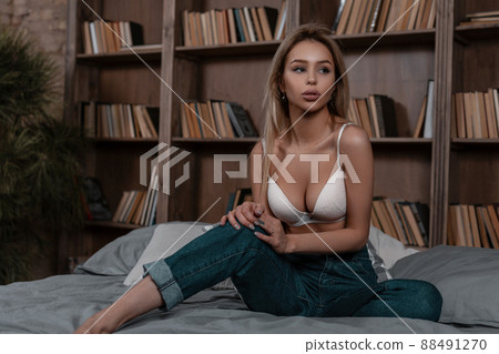 Gorgeous young blond woman in white elegant lingerie in fashionable vintage blue jeans resting and sitting on a bed indoors near the shelves of books. Attractive stylish girl model dreams. Gorgeous young blond woman in white elegant lingerie in fashionable vintage blue jeans resting and sitting on a bed indoors near the shelves of books. Attractive stylish girl model dreams. 88491270