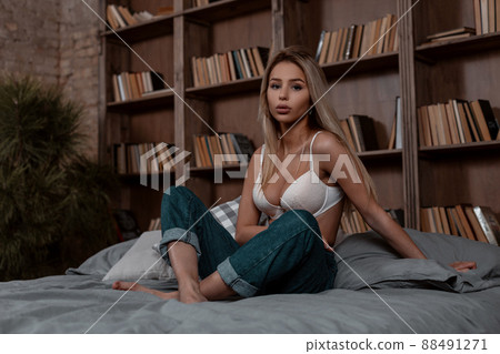 Cute sexy young blond woman in a white elegant bra in stylish blue jeans sitting on the bed in the bedroom. Pretty girl model is resting indoors near vintage wall with books. Cute sexy young blond woman in a white elegant bra in stylish blue jeans sitting on the bed in the bedroom. Pretty girl model is resting indoors near vintage wall with books. 88491271