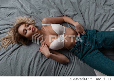 Attractive pretty stylish sexy slim young woman in a fashionable chic white bra with a beautiful lush breasts in stylish jeans is lying on the bed indoors. Top view of a glamorous girl. Attractive pretty stylish sexy slim young woman in a fashionable chic white bra with a beautiful lush breasts in stylish jeans is lying on the bed indoors. Top view of a glamorous girl. 88491275