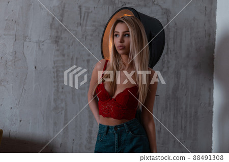 Young sexy pretty blonde in trendy fashion vintage blue jeans in a luxurious red lace bra is standing indoors near a metal light lamp. Sensual girl with a beautiful slim body. 88491308