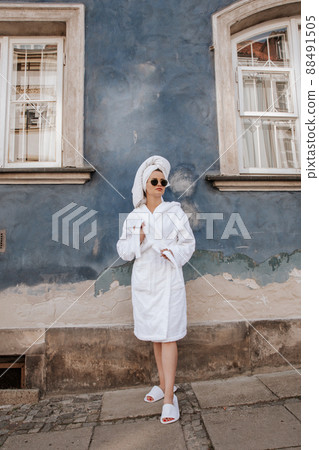Smiling cute girl blogger in white robe, towel and sunglasses stands on old street. Portrait young attractive model in a bathrobe, towel and sunglasses posing. Morning concept. 88491505