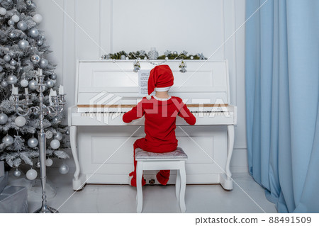 Boy in santa costume. Kid concept for merry Christmas and happy new year celebration. Small boy enjoys playing piano for the first time. Child play piano near the christmas tree. Christmas time. 88491509