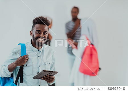 University lifestyle handsome young African student man holding a tablet computer and smiling while standing against university with his friends have a team meeting in the background. High-quality 88492948