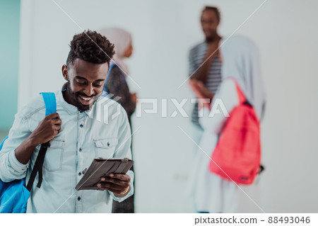 University lifestyle handsome young African student man holding a tablet computer and smiling while standing against university with his friends have a team meeting in the background. High-quality University lifestyle handsome young African student man holding a tablet computer and smiling while standing against university with his friends have a team meeting in the background. High-quality 88493046