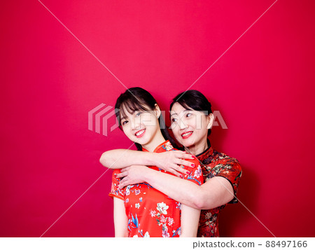Portrait of a daughter and mother wearing traditional cheongsam qipao and smiling cuddling isolated on red background. Portrait of a daughter and mother wearing traditional cheongsam qipao and smiling cuddling isolated on red background. 88497166