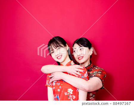 Portrait of a daughter and mother wearing traditional cheongsam qipao and smiling cuddling isolated on red background. 88497167