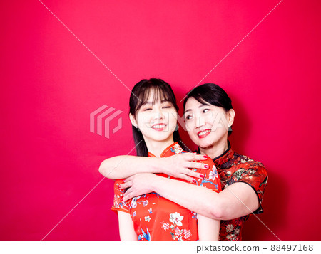 Portrait of a daughter and mother wearing traditional cheongsam qipao and smiling cuddling isolated on red background. Portrait of a daughter and mother wearing traditional cheongsam qipao and smiling cuddling isolated on red background. 88497168