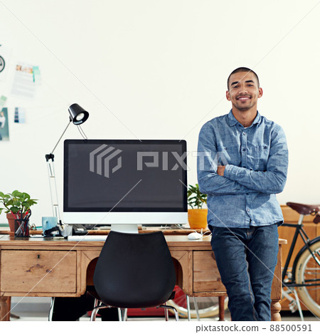 He brings designs to life. Portrait of a casually-dressed young man sitting on his desk in an office. He brings designs to life. Portrait of a casually-dressed young man sitting on his desk in an office. 88500591