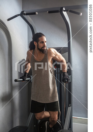 Full length portrait of confident young adult strong man with long curly hair working out in gym, hanging on horizontal bar with hands, doing pull ups with, exercises for triceps. indoor, looking away 88501086