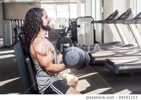 Side view portrait of concentration young adult man with long curly hair working out in gym, sitting on bench and holding two dumbbell with raised arms, doing exercises for biceps. indoor,looking away Side view portrait of concentration young adult man with long curly hair working out in gym, sitting on bench and holding two dumbbell with raised arms, doing exercises for biceps. indoor,looking away 88501153