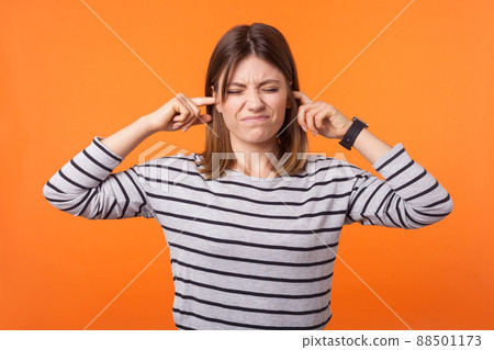 Don't want to hear it. Portrait of irritated woman with brown hair in long sleeve shirt standing grimacing and closing ears from noise, avoid conflict. indoor studio shot isolated on orange background Don't want to hear it. Portrait of irritated woman with brown hair in long sleeve shirt standing grimacing and closing ears from noise, avoid conflict. indoor studio shot isolated on orange background 88501173