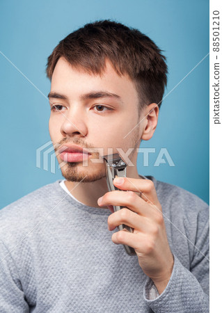 Grooming and self care. Portrait of concentrated focused brunette man shaving with trimmer or electric shaver, looking aside with serious attentive face. studio shot isolated on blue background Grooming and self care. Portrait of concentrated focused brunette man shaving with trimmer or electric shaver, looking aside with serious attentive face. studio shot isolated on blue background 88501210