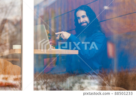 Portrait through the window of cheerful young adult man freelancer in casual style sitting in cafe, talking with friend in laptop, pointing finger to screen, choosing you. outdoor, lifestyle concept Portrait through the window of cheerful young adult man freelancer in casual style sitting in cafe, talking with friend in laptop, pointing finger to screen, choosing you. outdoor, lifestyle concept 88501233
