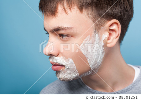 Closeup profile of young confident man looking aside with shaving foam on face, showing smooth soft skin on shaved part of cheek, moisturizing cosmetics. indoor studio shot isolated on blue background 88501251