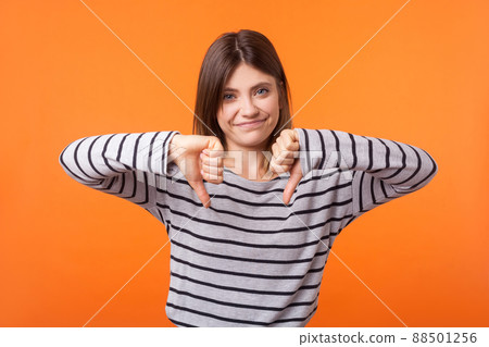 Portrait of dissatisfied young woman with brown hair in long sleeve striped shirt standing, showing disapproval and rejection with thumbs down gesture. indoor studio shot isolated on orange background 88501256