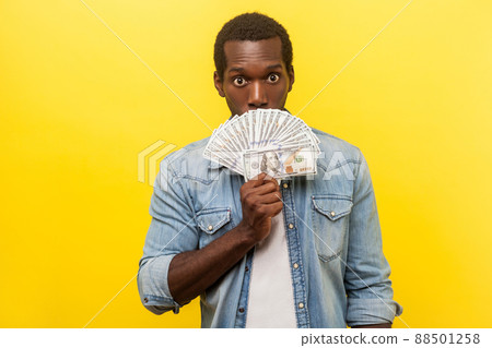 Portrait of surprised scared man in denim casual shirt holding dollar bills, peeking out of money while hiding half face with astonished shocked eyes. indoor studio shot isolated on yellow background 88501258