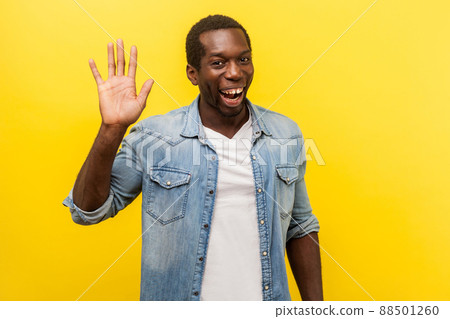 Hello. Portrait of positive handsome man in denim casual shirt with rolled up sleeves smiling friendly and waving hand saying hi, welcoming gesture. indoor studio shot isolated on yellow background Hello. Portrait of positive handsome man in denim casual shirt with rolled up sleeves smiling friendly and waving hand saying hi, welcoming gesture. indoor studio shot isolated on yellow background 88501260