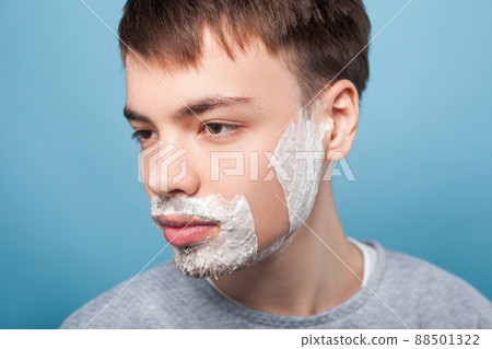 Portrait of serious man in casual clothes looking aside with shaving foam on his bristle and mustache, showing smooth soft skin on shaved part of cheek. indoor studio shot isolated on blue background Portrait of serious man in casual clothes looking aside with shaving foam on his bristle and mustache, showing smooth soft skin on shaved part of cheek. indoor studio shot isolated on blue background 88501322
