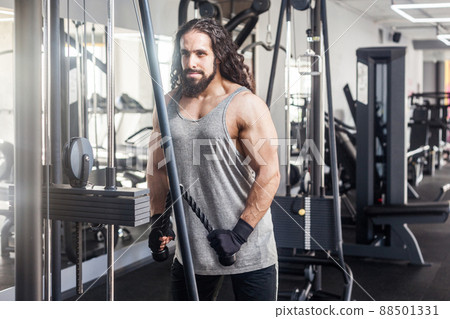 Portrait of young adult sport athlete man with long curly hair training at gym alone, standing and lifting weights in the gym, doing exercises for triceps. Healthy lifestyle concept, indoor 88501331
