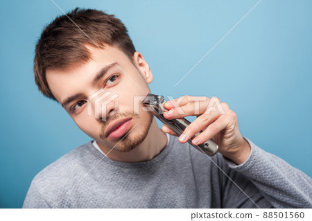 Shaving routine. Portrait of young handsome brunette man in casual sweater shaving with trimmer looking up as if in mirror to shape his beard, attentive and serious. studio shot isolated on blue Shaving routine. Portrait of young handsome brunette man in casual sweater shaving with trimmer looking up as if in mirror to shape his beard, attentive and serious. studio shot isolated on blue 88501560