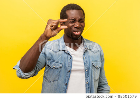 Pinching hand gesture. Portrait of positive man in denim shirt showing a little bit sign with fingers, making imploring grimace, need some more. indoor studio shot isolated on yellow background 88501579