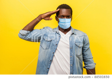 Yes sir. Portrait of serious confident man in denim casual shirt with surgical medical mask standing as soldier giving salute, ready for order. indoor studio shot isolated on yellow background 88501650