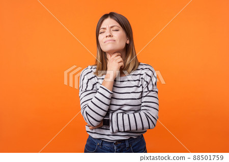 Portrait of ill woman with brown hair in long sleeve striped shirt standing touching neck and frowning from pain, suffering sore throat, flu symptom. indoor studio shot isolated on orange background Portrait of ill woman with brown hair in long sleeve striped shirt standing touching neck and frowning from pain, suffering sore throat, flu symptom. indoor studio shot isolated on orange background 88501759