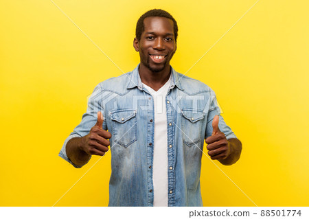 Thumbs up, excellent job. Portrait of enthusiastic handsome man in denim casual shirt with rolled up sleeves smiling and showing like gesture at camera. studio shot isolated on yellow background 88501774