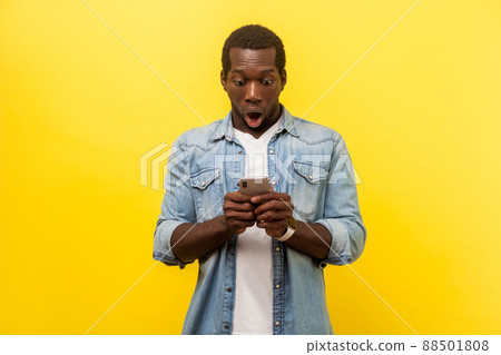 Portrait of amazed man in denim casual shirt using cellphone, typing text message or dialing number with shocked face expression, technology concept. indoor studio shot isolated on yellow background 88501808