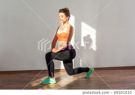 Split or lung squats. Portrait of fit woman with bun hairstyle and in tight sportswear practicing with her eyes closed, lower-body exercise. indoor studio shot illuminated by sunlight from window 88501810