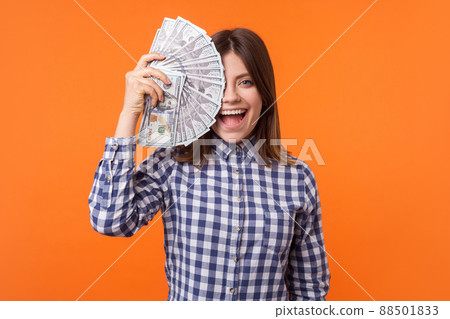 Half face portrait of amazed attractive brunette woman wearing checkered shirt holding dollars and looking at camera with open mouth, lottery winner. indoor studio shot isolated on orange background 88501833