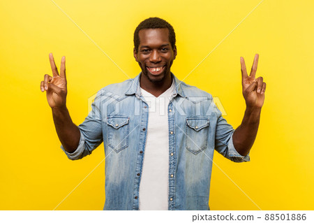Successful winner. Portrait of excited man with toothy smile in denim casual shirt standing with raised hands and showing v sign or peace, victory gesture. studio shot isolated on yellow background 88501886