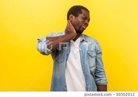 Portrait of unhealthy tired man in denim casual shirt with rolled up sleeves touching neck suffering pain, muscle tension or injury, overwork concept. indoor studio shot isolated on yellow background Portrait of unhealthy tired man in denim casual shirt with rolled up sleeves touching neck suffering pain, muscle tension or injury, overwork concept. indoor studio shot isolated on yellow background 88502012