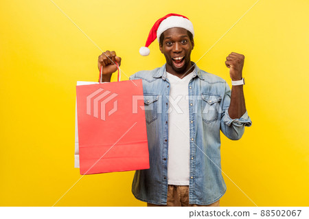 Portrait of amazed happy man in santa hat and casual denim shirt standing with raised hand showing yes gesture, holding bags, excited about shopping. indoor studio shot isolated on yellow background Portrait of amazed happy man in santa hat and casual denim shirt standing with raised hand showing yes gesture, holding bags, excited about shopping. indoor studio shot isolated on yellow background 88502067