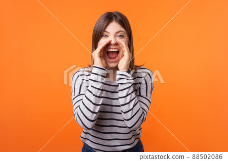 Attention. Portrait of stressed furious young woman with brown hair in long sleeve shirt standing, holding arms near wide open mouth and screaming. indoor studio shot isolated on orange background 88502086