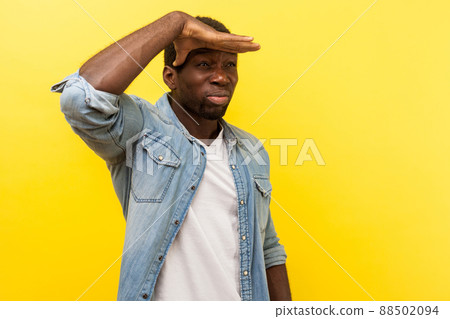 Portrait of concentrated attentive man in denim casual shirt with rolled up sleeves holding hand over eyes and looking at long distance, far future. indoor studio shot isolated on yellow background 88502094