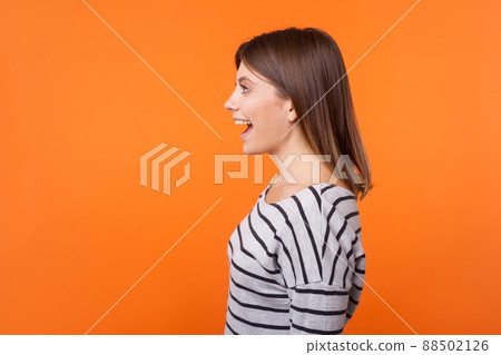 Side view portrait of beautiful amazed woman with brown hair in long sleeve striped shirt standing surprised with open mouth, looking to the left. indoor studio shot isolated on orange background 88502126