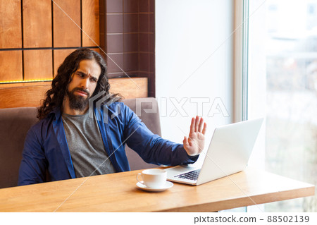 Serious handsome young adult man freelancer in casual style sitting in cafe and talking with his opponent in laptop, showing hand to notebook like to stop. Indoor, lifestyle concept Serious handsome young adult man freelancer in casual style sitting in cafe and talking with his opponent in laptop, showing hand to notebook like to stop. Indoor, lifestyle concept 88502139