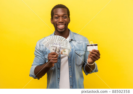 Portrait of excited young man in denim shirt holding dollar bills and credit card with satisfied expression, toothy smile, enjoying payments with cashback. studio shot isolated on yellow background Portrait of excited young man in denim shirt holding dollar bills and credit card with satisfied expression, toothy smile, enjoying payments with cashback. studio shot isolated on yellow background 88502358