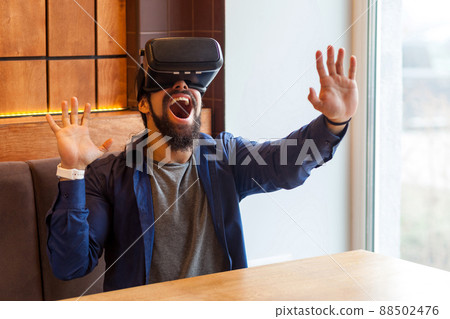 Portrait of happy unbelievable bearded young adult man in casual style sitting in cafe, wearing vr and watching video on simulator with amazing happy face and raised arms. Indoor, lifestyle Portrait of happy unbelievable bearded young adult man in casual style sitting in cafe, wearing vr and watching video on simulator with amazing happy face and raised arms. Indoor, lifestyle 88502476