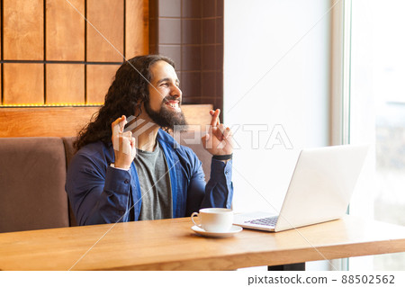 Portrait of dreaming handsome young adult man freelancer in casual style sitting in cafe with laptop, showing crossed finger with closed eyes, toothy smile, bussinessman in office. Indoor, lifestyle 88502562