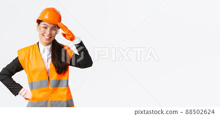 Confident smiling, friendly asian female construction manager, engineer in safety helmet and gloves saluting, greeting architect or chief at enterprise, showing building area to investors 88502624