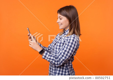 Side view portrait of adorable young brunette woman with charming smile wearing checkered shirt standing with cellphone, making video call or blogging. indoor studio shot isolated on orange background Side view portrait of adorable young brunette woman with charming smile wearing checkered shirt standing with cellphone, making video call or blogging. indoor studio shot isolated on orange background 88502751