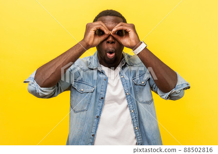 Portrait of astonished young man in denim casual shirt looking at camera through fingers in binoculars gesture with surprised expression, saying wow. indoor studio shot isolated on yellow background 88502816