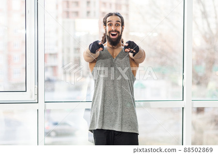 Portrait of suprised happy young adult muscular trainer man with curly long hair standing, pointing fingers to you with big eyes, looking at camera with toothy smile. Indoor, window background 88502869