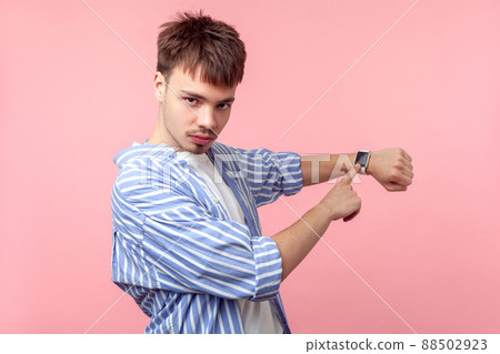 Look at the time. Portrait of irritated brown-haired man with small beard and mustache in casual shirt looking angrily and pointing at his wristwatch. indoor studio shot isolated on pink background Look at the time. Portrait of irritated brown-haired man with small beard and mustache in casual shirt looking angrily and pointing at his wristwatch. indoor studio shot isolated on pink background 88502923